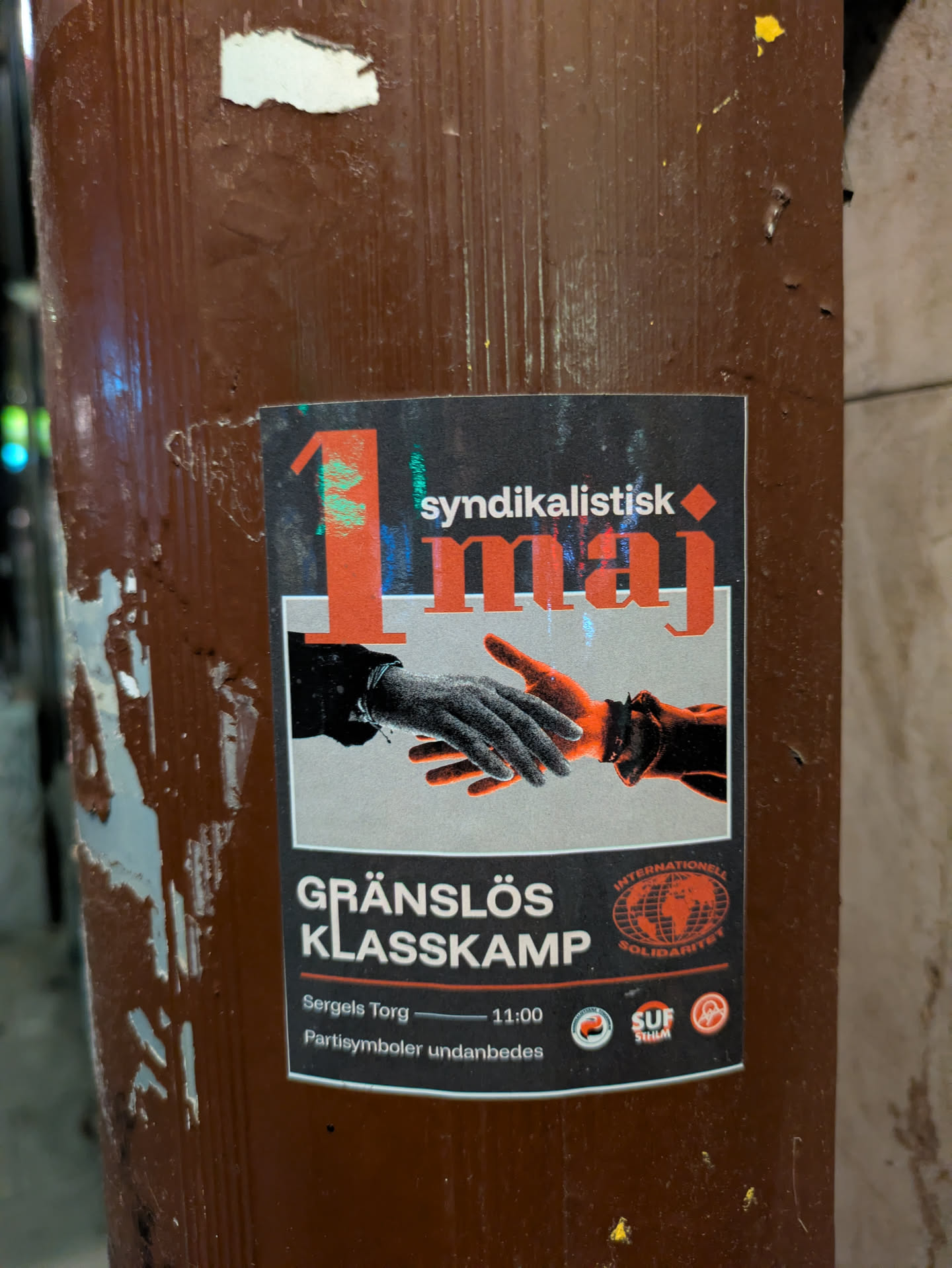 The sticker features the text "Syndikalistisk 1 maj" with an image of two hands shaking, symbolizing unity. Below the image, the sticker continues with "Gränslös Klasskamp" and includes details about a gathering at Sergels Torg at 11:00. It also contains the phrase "Partisymboler undanbedes", which roughly translates to 'Party symbols not desired'. The sticker has logos for the SAC (Syndicalistiska Arbetarekommunen) and SUF (Syndicalist Youth Federation) as well as the International Solidarity logo.