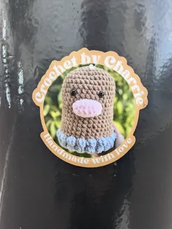 Street sticker A round sticker featuring a crocheted amigurumi toy. The sticker has a beige background with brown text that reads: 'Crochet by Charlie' in a circular design. Inside the circle is a picture of a brown crocheted amigurumi toy with a pink nose and light blue accents. Below the toy, the text 'Handmade with love' is written.
