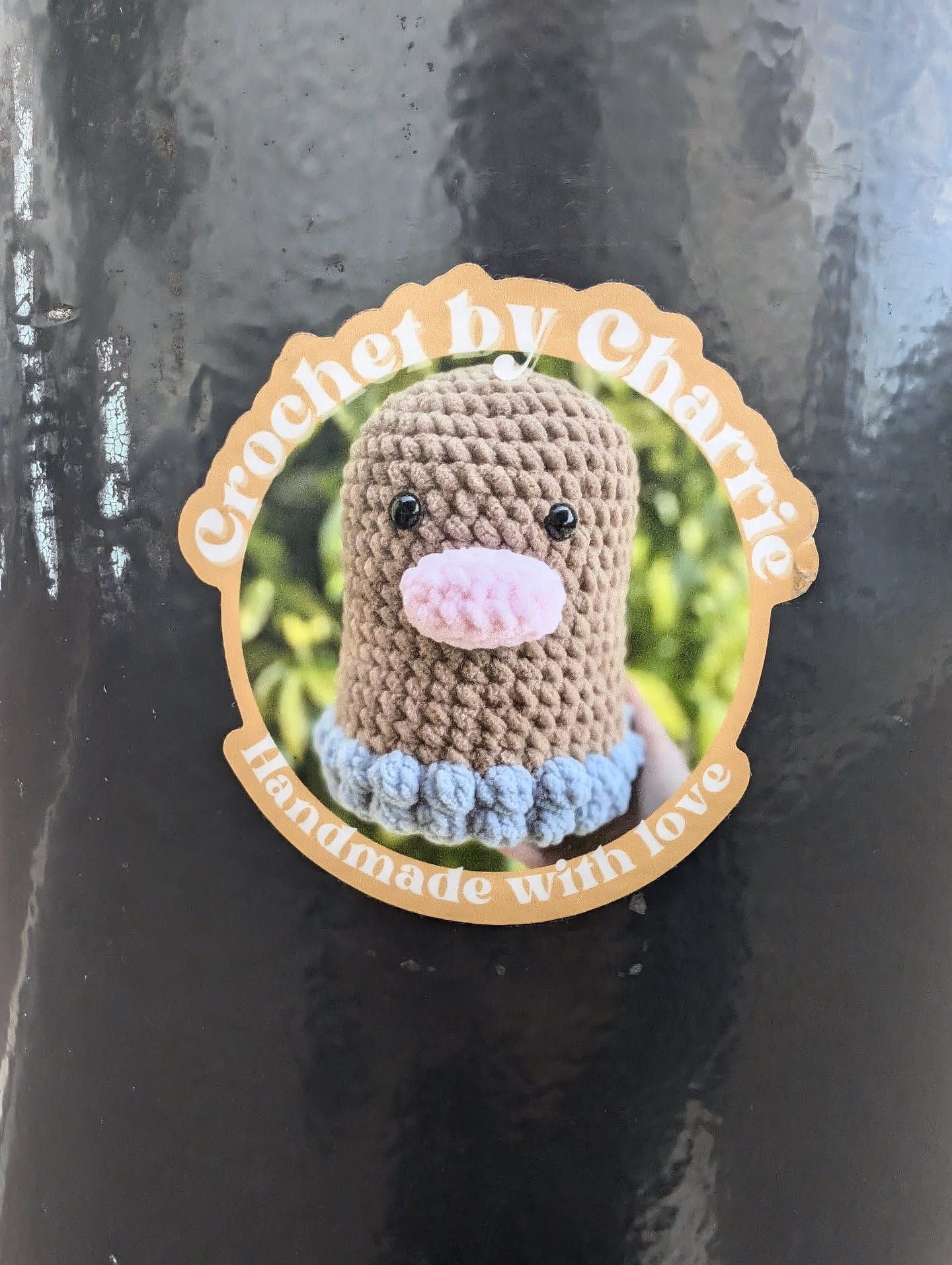 A round sticker featuring a crocheted amigurumi toy. The sticker has a beige background with brown text that reads: 'Crochet by Charlie' in a circular design. Inside the circle is a picture of a brown crocheted amigurumi toy with a pink nose and light blue accents. Below the toy, the text 'Handmade with love' is written.
