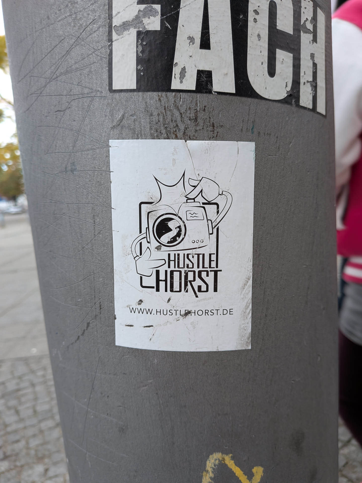 A vertical rectangular sticker on a metal post. Printed in black on a white background, the central graphic shows an anthropomorphic vintage-style camera character. The character has cartoon arms: one hand points forward like a finger gun, while the other arm loops over the top to press the shutter button, creating a stylized flash burst above it. Beneath the illustration, the bold stylized text reads 'HUSTLE HORST', framed in black line borders, with the website 'WWW.HUSTLEHORST.DE' printed at the bottom.