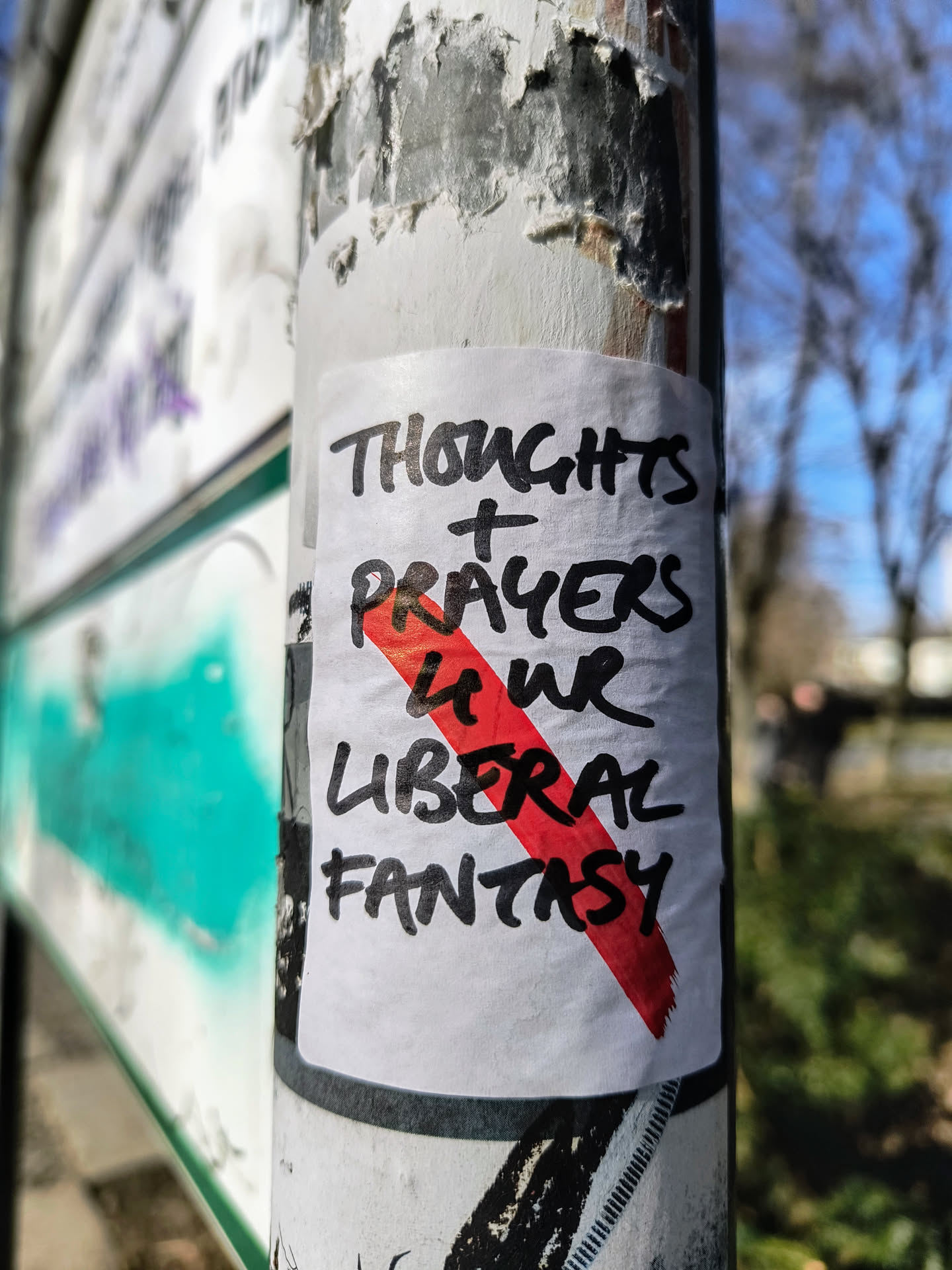 A white square sticker on a pole with the text "Thoughts + Prayers" written in bold black marker, with the words "Your Liberal Fantasy" written below. A thick red line crosses out "Your Liberal Fantasy". The pole is dirty and covered in other scraps of paper.