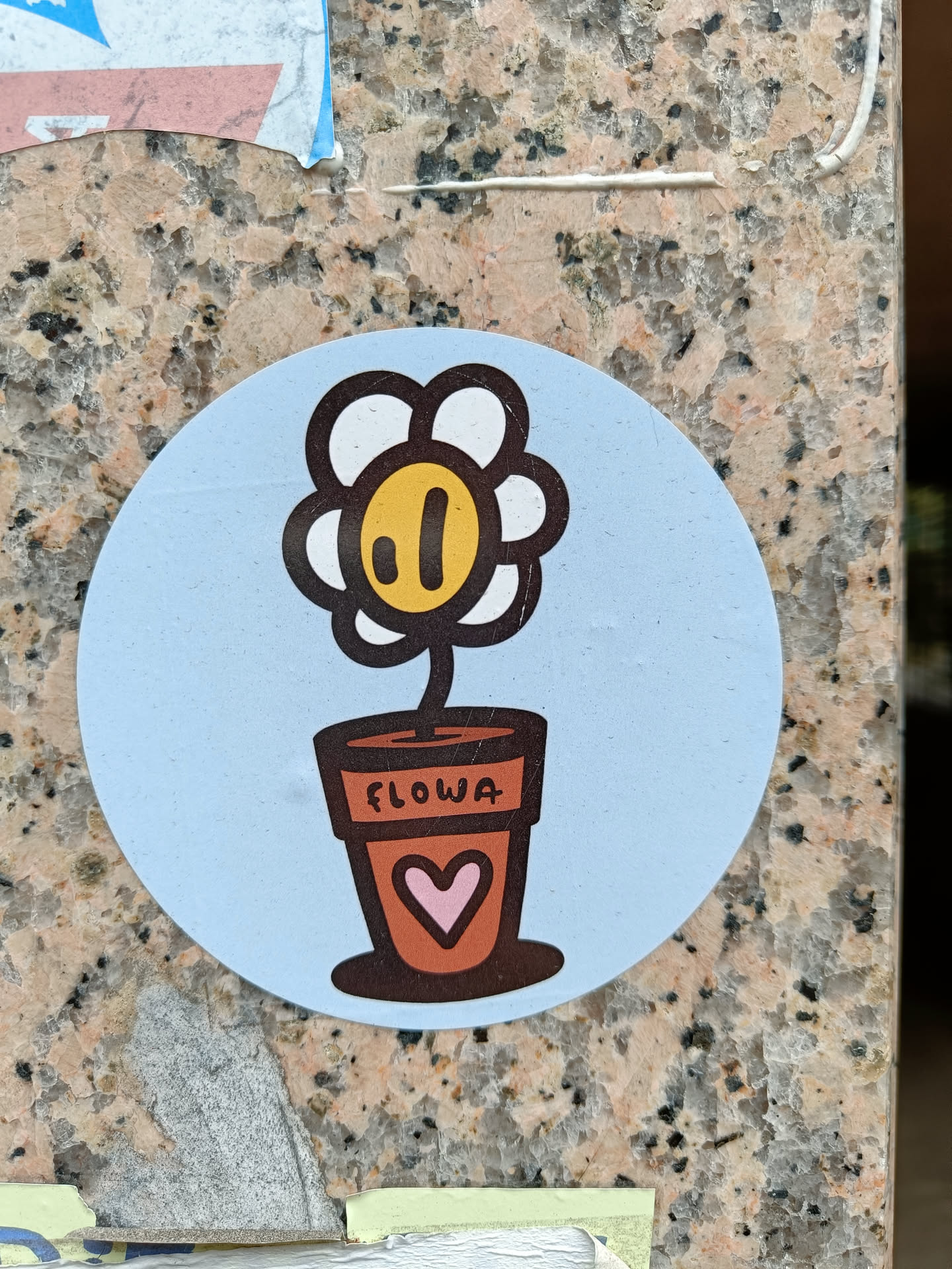 A round sticker with a light blue background features a simple drawing of a flower in a pot. The flower is white with a yellow center and a brown stem. The pot is brown with the word "FLOWA" written on it and a small pink heart.