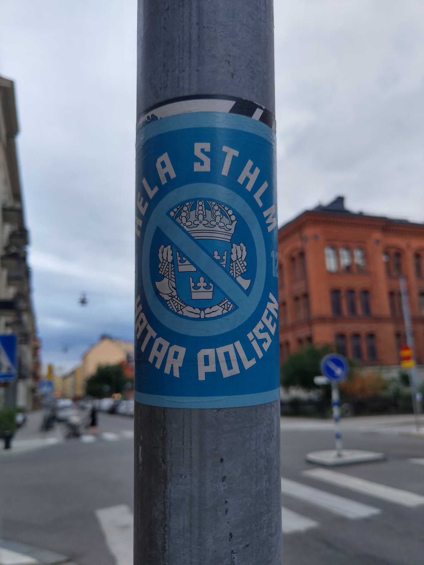 A blue and white sticker affixed to a lamppost. The sticker features a circular design with a crossed-out coat of arms and text that appears to say "Hela Sthlm hatar polisen".  The sticker is in Swedish.