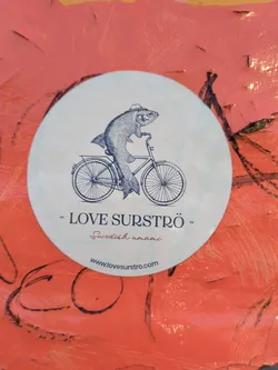 Oval sticker with a line drawing of a fish wearing a hat and riding a bicycle.  Text on the sticker says "LOVE SURSTRÖM" and "Swedish umami" with a website address at the bottom.