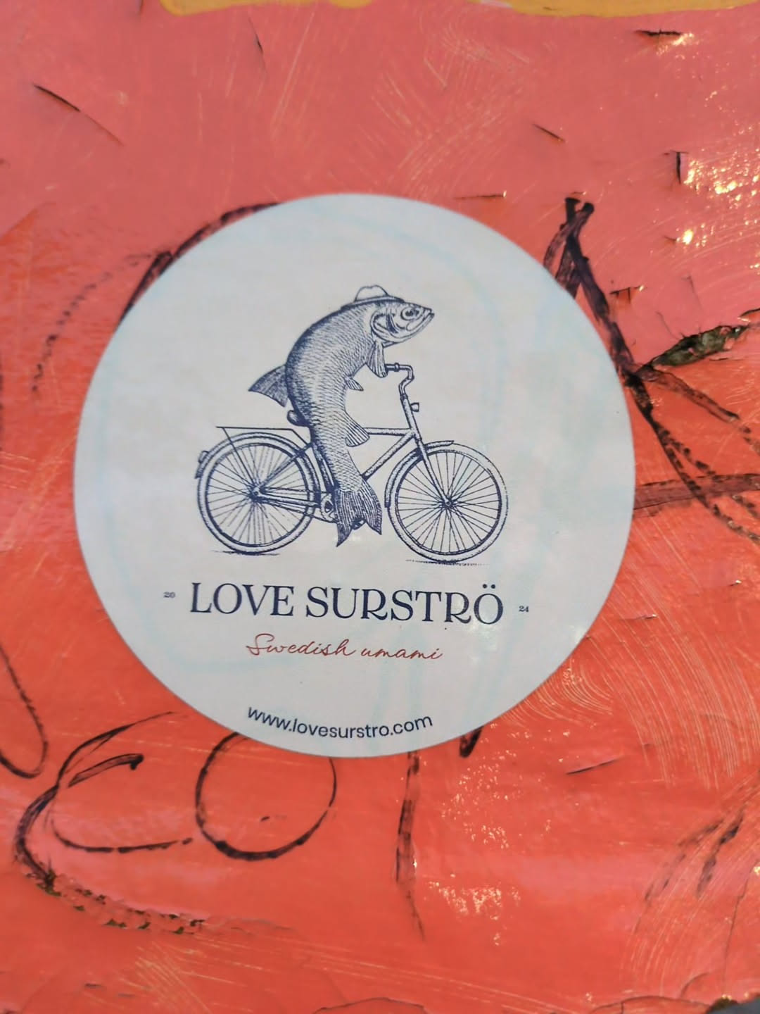 Oval sticker with a line drawing of a fish wearing a hat and riding a bicycle.  Text on the sticker says "LOVE SURSTRÖM" and "Swedish umami" with a website address at the bottom.