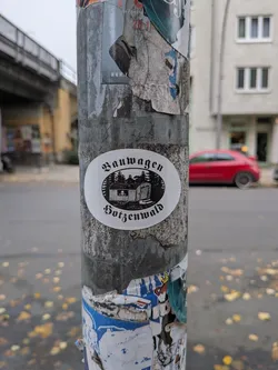 Street sticker An oval-shaped white sticker with black ink printing. In the center is a woodcut-style illustration of a traditional 'Bauwagen' (mobile construction trailer or living wagon) situated in front of a pine forest under a clear sky, framed within a horizontal oval shape. Arching above the illustration is the German word 'Bauwagen' and arching below is 'Hotzenwald', both written in an ornate Fraktur / Blackletter typeface.