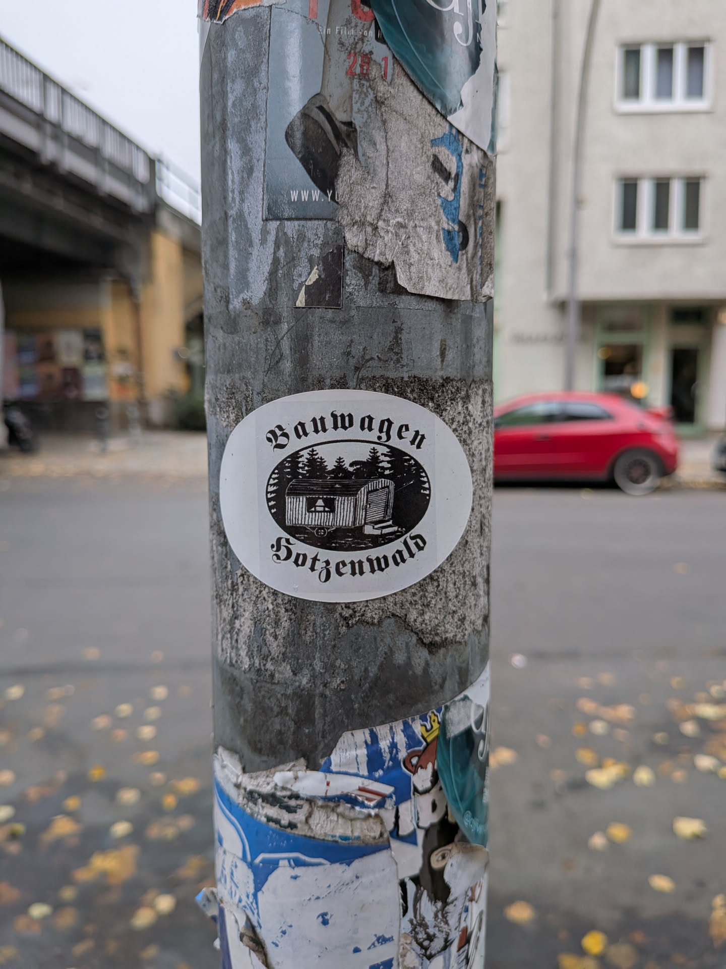 An oval-shaped white sticker with black ink printing. In the center is a woodcut-style illustration of a traditional 'Bauwagen' (mobile construction trailer or living wagon) situated in front of a pine forest under a clear sky, framed within a horizontal oval shape. Arching above the illustration is the German word 'Bauwagen' and arching below is 'Hotzenwald', both written in an ornate Fraktur / Blackletter typeface.