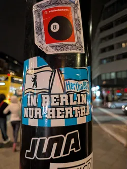 A rectangular sticker adhering to a metal pole, featuring light cyan-blue and white vertical stripes on the background. In the upper portion, two waving flags are displayed: the left flag depicts the Berlin bear silhouette, and the right flag contains the text 'Hertha BSC'. Centered between the flags is a stylized outline of the Kaiser Wilhelm Memorial Church spire. Below the flags, bold white capital letters outlined in black declare 'IN BERLIN NUR HERTHA'. The top edge of the sticker shows some slight tearing and wear.