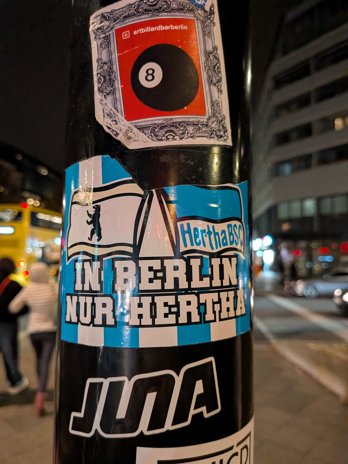 A rectangular sticker adhering to a metal pole, featuring light cyan-blue and white vertical stripes on the background. In the upper portion, two waving flags are displayed: the left flag depicts the Berlin bear silhouette, and the right flag contains the text 'Hertha BSC'. Centered between the flags is a stylized outline of the Kaiser Wilhelm Memorial Church spire. Below the flags, bold white capital letters outlined in black declare 'IN BERLIN NUR HERTHA'. The top edge of the sticker shows some slight tearing and wear.