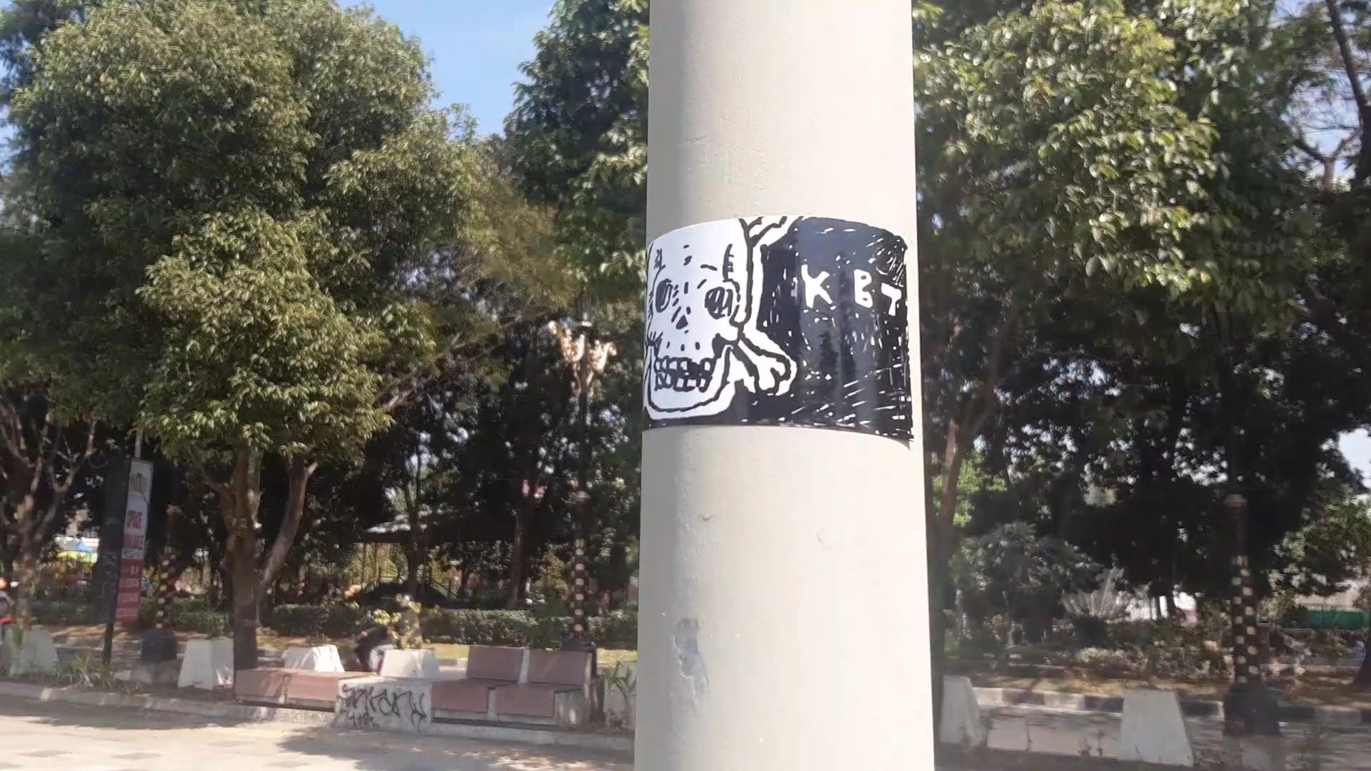 A rectangular monochrome sticker affixed to a light-colored outdoor pole. The design features a hand-drawn skull with a bone cross motif on the left side over a white area, contrasting with a heavily shaded black background on the right. Scrawled in rough white lettering within the dark right section is the text 'KBT'.