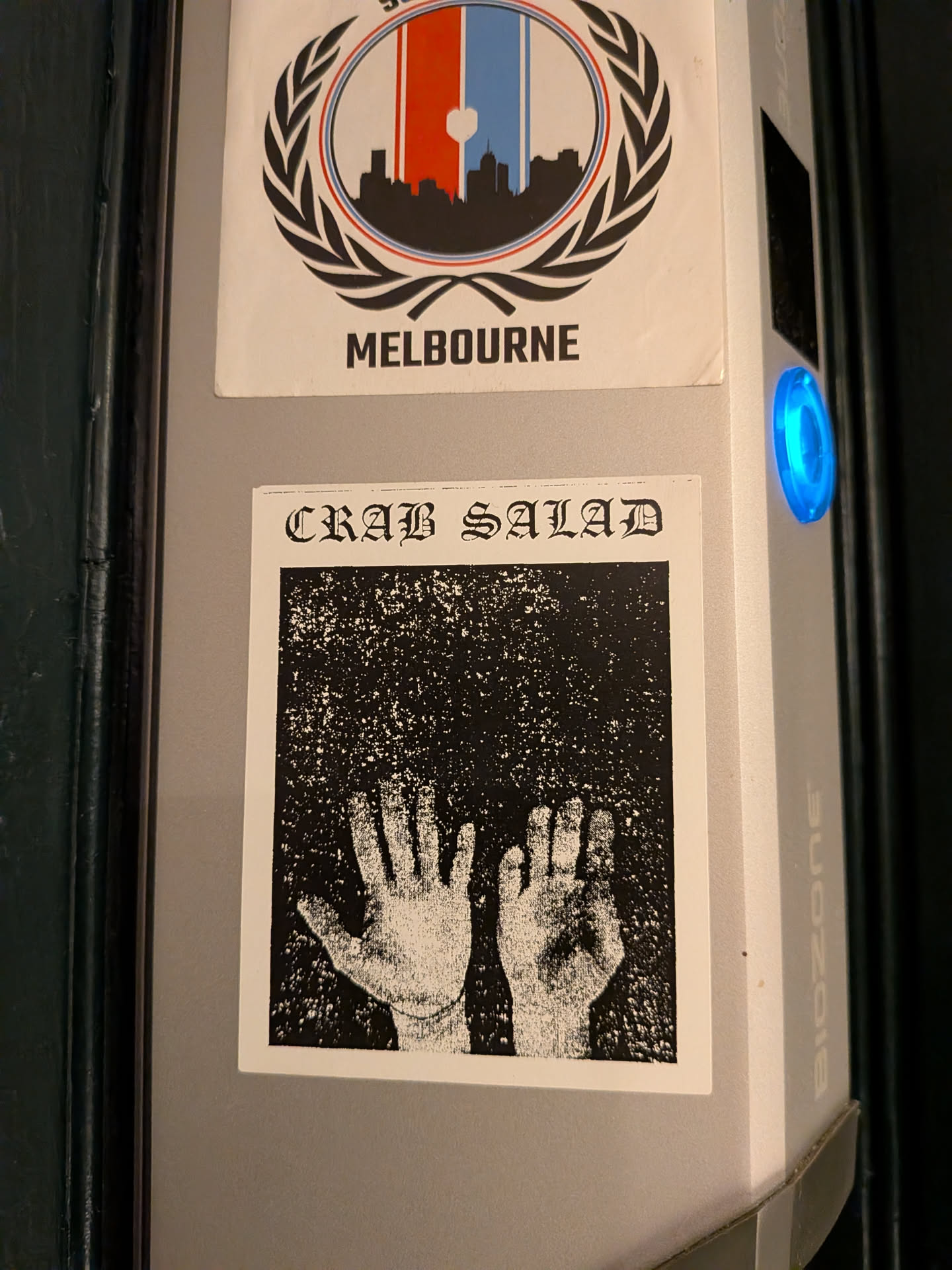 A square, black and white sticker with a thin white border. The top features the text 'CRAB SALAD' in a distinctive, gothic-style font. Below the text, two hands are depicted, palms facing outwards with fingers slightly spread, rendered in a highly distressed, grainy, and high-contrast style. The hands appear light against a dark, speckled background, giving them an ethereal or ghostly quality. The overall aesthetic is raw, gritty, and punk-inspired.