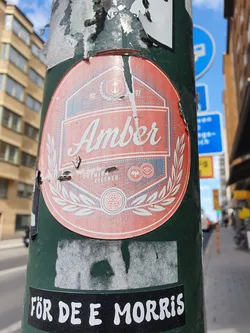 A sticker featuring a vintage-style design for Amber Premium Pilsner. The sticker is peach-colored with a navy blue and white emblem.  It's affixed to a green pole, partially obscured by other stickers and showing some wear and tear.