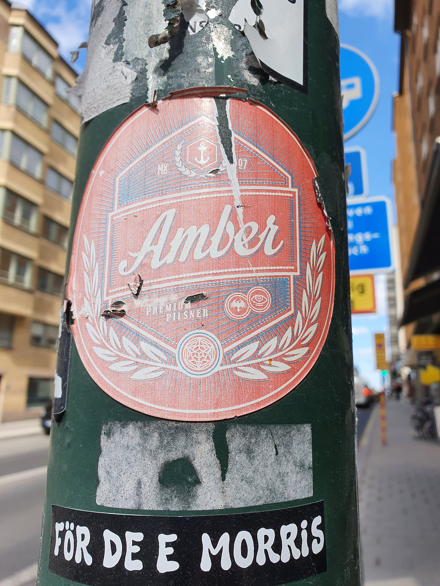 A sticker featuring a vintage-style design for Amber Premium Pilsner. The sticker is peach-colored with a navy blue and white emblem.  It's affixed to a green pole, partially obscured by other stickers and showing some wear and tear.