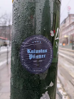 A dark blue and light blue circular sticker featuring the text "Knästas Pilsner" in a gothic-style font.  The sticker also includes the years "1932 - 2008" and the text "Sandbergs Magiska" in a smaller font. The sticker is affixed to a dark green pole covered in water droplets, suggesting a damp or cold environment. The background shows a blurry city street scene.