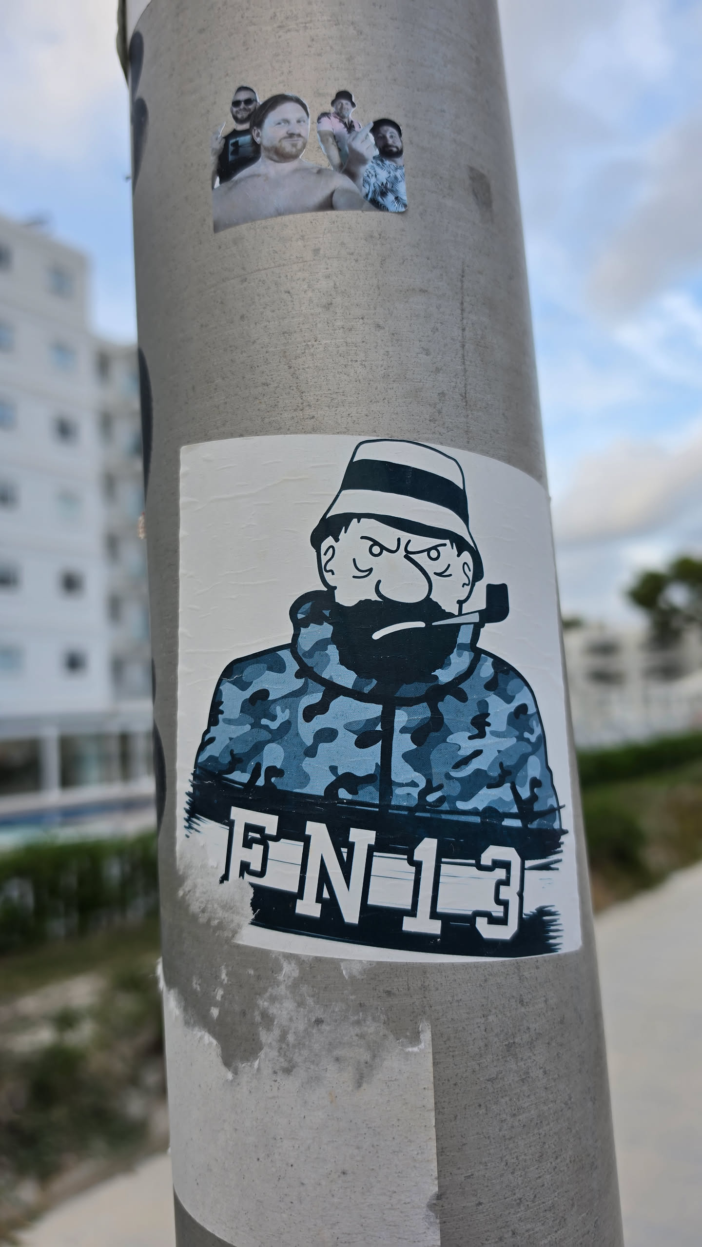 A rectangular sticker on a metal pole featuring a stylized cartoon character resembling Captain Haddock. The character has an angry facial expression, a full black beard, a pipe clenched in his teeth, and is wearing a bucket hat with a band and a blue-and-gray camouflage hooded jacket. Below the figure, bold block text reads 'FN13' displayed across a dark horizontal banner.
