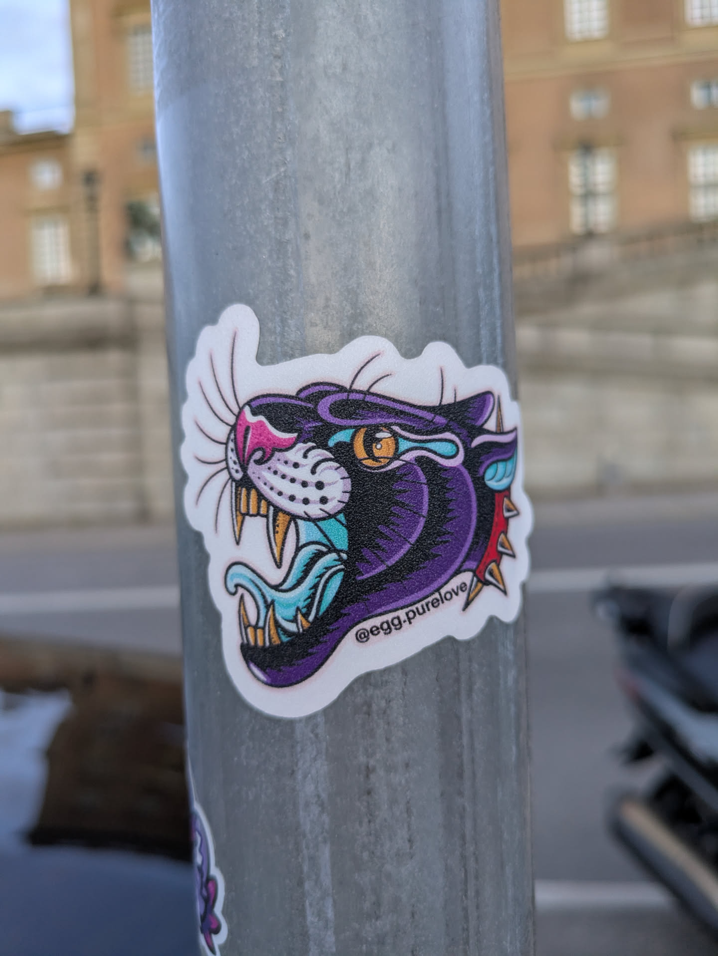 The sticker features the head of a black panther in profile, rendered in a bold, tattoo-inspired style. The panther's eyes are a striking yellow with black pupils, outlined in purple. Its mouth is open, revealing sharp, yellow fangs and a blue and white tongue. Accents of pink are visible on its nose and inner ears. The creature is depicted with a vibrant purple mane or fur, highlighted with black shading for depth. Around its neck, a collar-like element with sharp, red and yellow spikes is visible. The overall color palette is rich and contrasting, featuring purple, black, teal, pink, yellow, and red. The sticker has a white border and a textured appearance, suggesting it's made of vinyl.