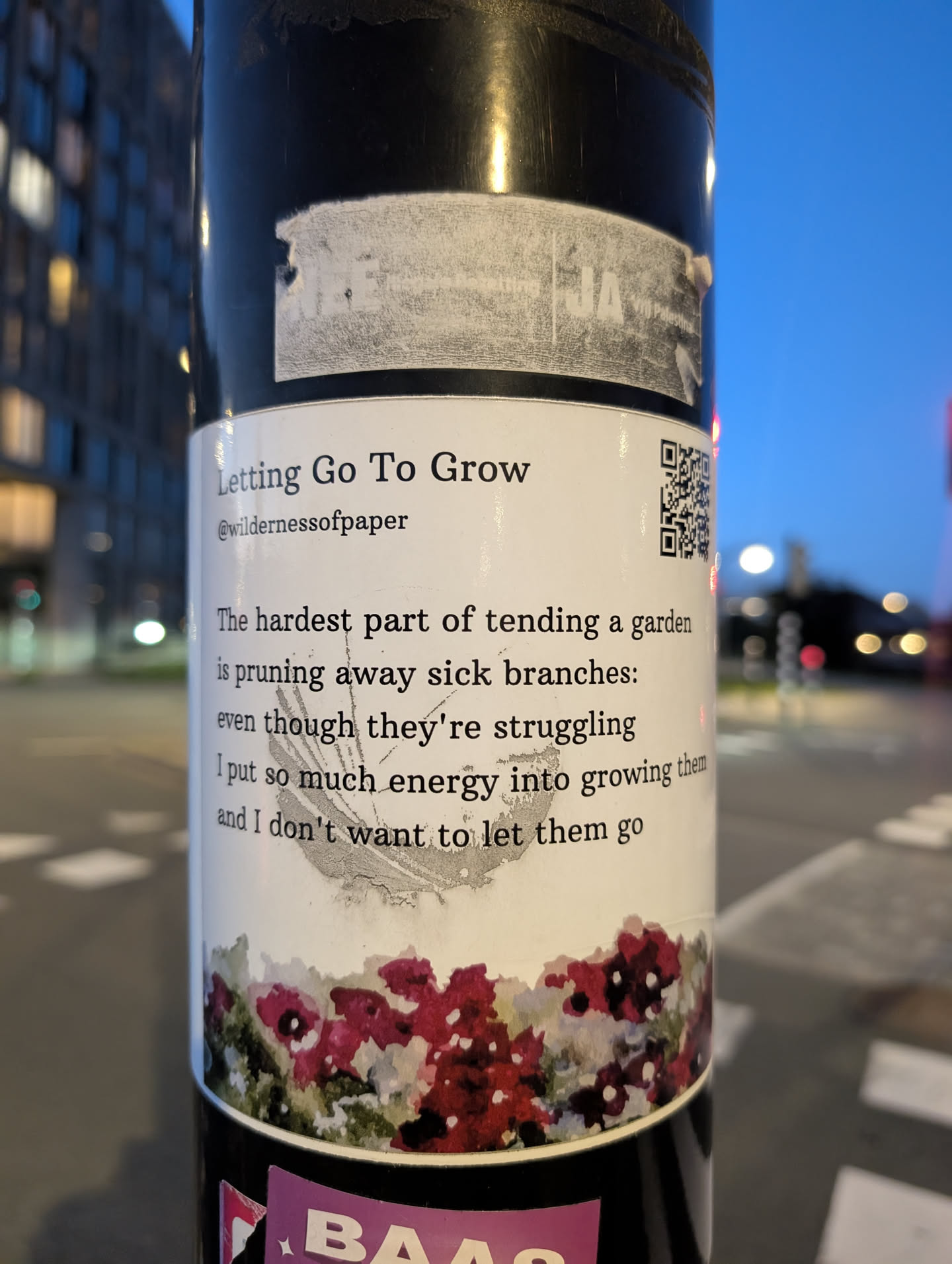 The sticker is predominantly white with black text. At the top, it reads 'Letting Go To Grow' in a serif font, followed by a social media handle '@wildernessofpaper'. Below this, a paragraph of text discusses the difficulty of pruning sick branches in a garden, relating it to the energy invested in things that are struggling and the reluctance to let them go. The text is accompanied by a small, stylized illustration of dark pink and red flowers with green foliage at the bottom. To the right of the text, a QR code is visible. The sticker appears to be affixed to a dark, cylindrical object, possibly a pole.
