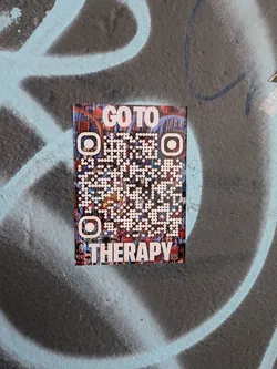 Street sticker A vertical rectangular sticker affixed to a textured, spray-painted surface. The sticker has an abstract, painterly backdrop composed of rich reds, blues, and dark tones. Overlaid on this art is a stylized QR code composed of white circular dots and custom rounded finder patterns, framed by bold white sans-serif text reading 'GO TO' at the top and 'THERAPY' across the bottom.