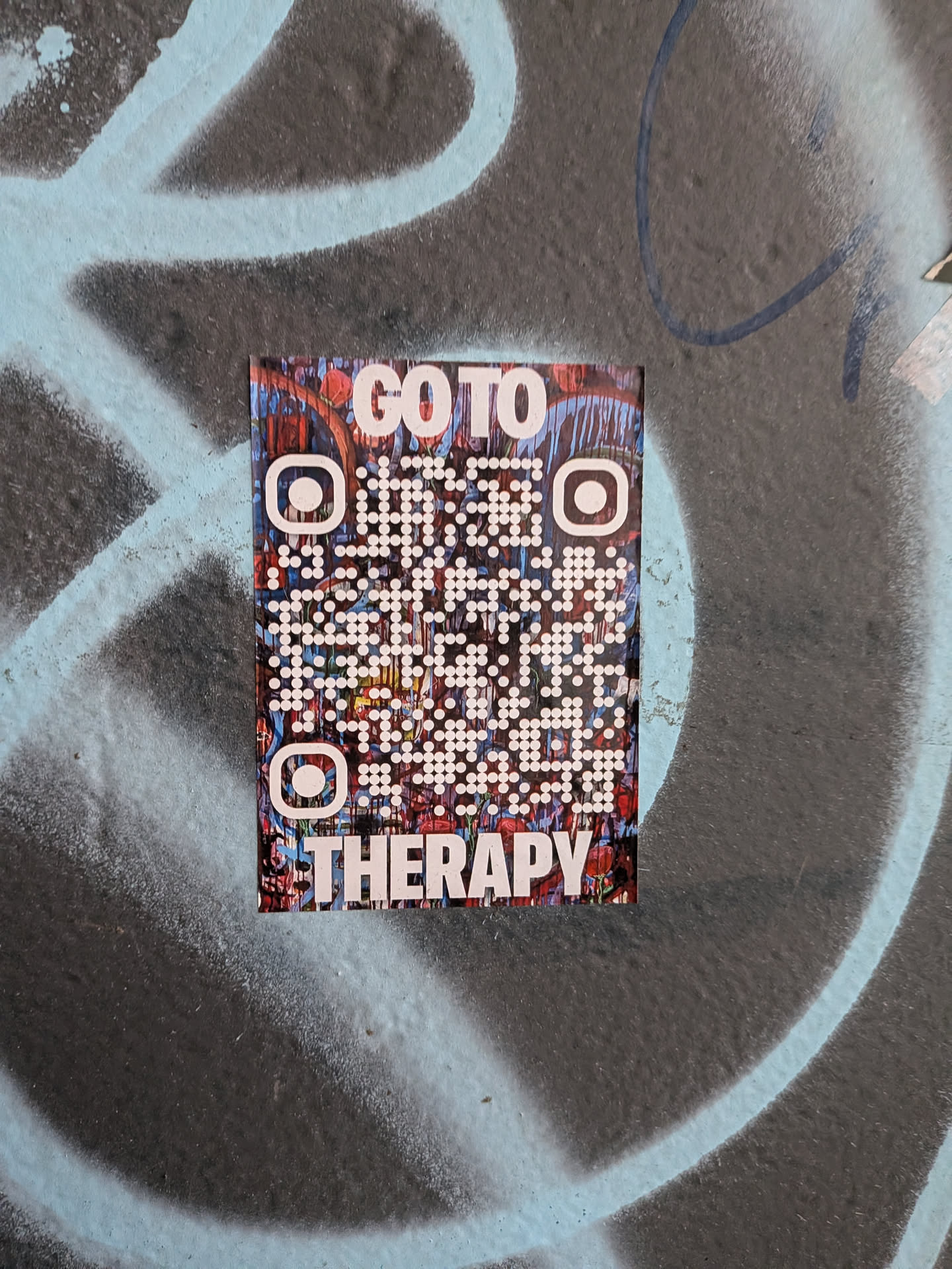 A vertical rectangular sticker affixed to a textured, spray-painted surface. The sticker has an abstract, painterly backdrop composed of rich reds, blues, and dark tones. Overlaid on this art is a stylized QR code composed of white circular dots and custom rounded finder patterns, framed by bold white sans-serif text reading 'GO TO' at the top and 'THERAPY' across the bottom.