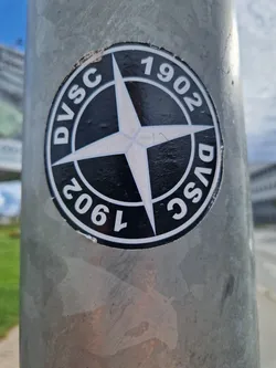 A black and white circular sticker with a compass rose and the text DVSC 1902 repeated. It's affixed to a gray metal pole.