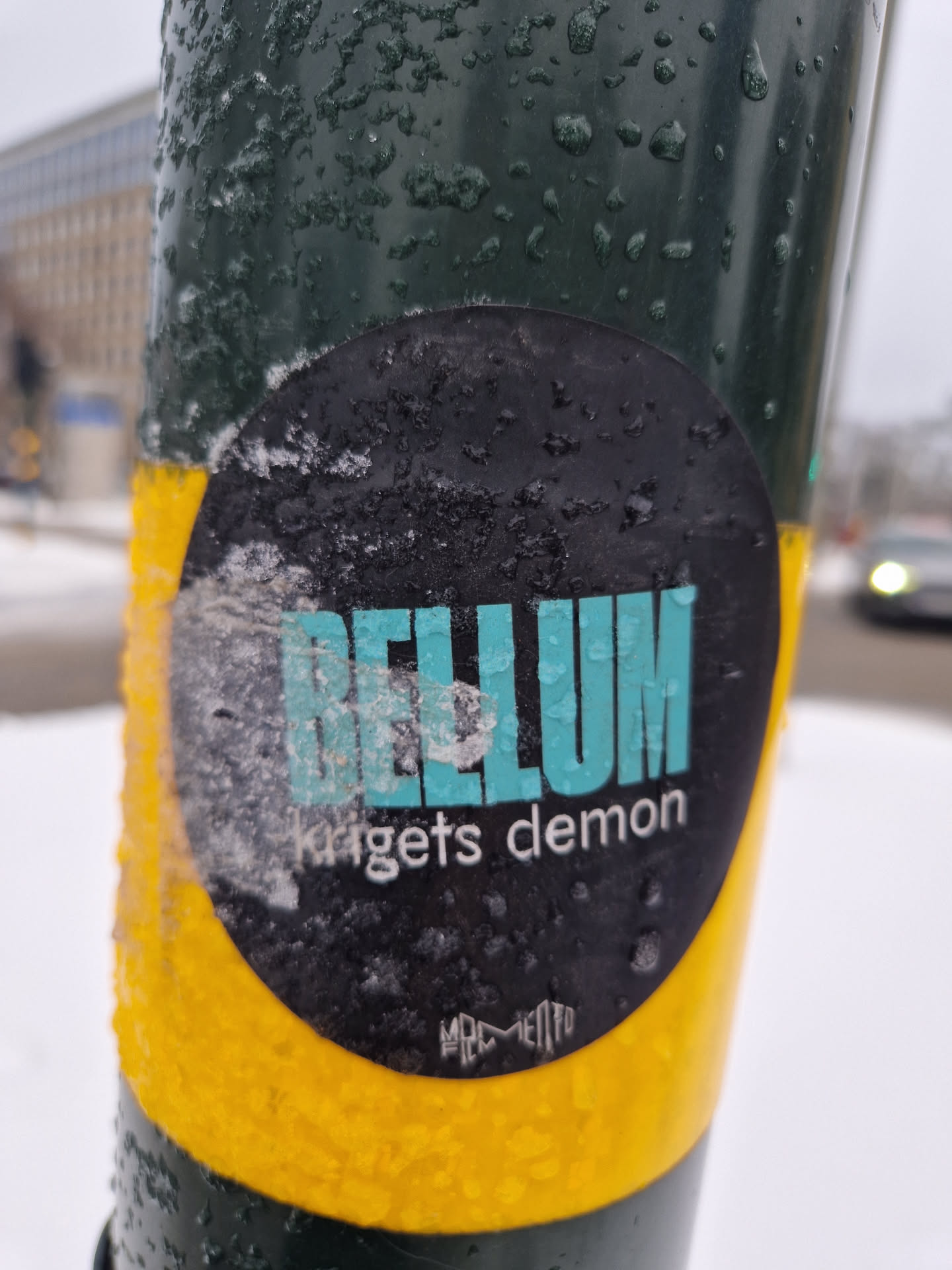 A round sticker with the text "BELLUM" in teal and "krigets demon" in white underneath. The sticker has a black background and a yellow border. It is attached to a dark green pole covered in ice or snow.