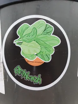 Round sticker featuring an illustration of spinach on a black background. The word 'SPINACH' is written in green dripping letters below the illustration.  The artist's Instagram handle is visible on the sticker.