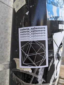 This sticker on a pole shows the username "materia_ephemera" repeated and a geometric design with a faint text saying "imagine yourself".