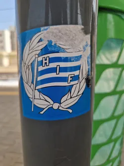 A blue sticker featuring a stylized flag with the letters HIF on it, surrounded by a laurel wreath. The sticker is affixed to a grey pole. The flag is blue and white striped, with the letters in a sans-serif font. The sticker shows some signs of wear and tear.