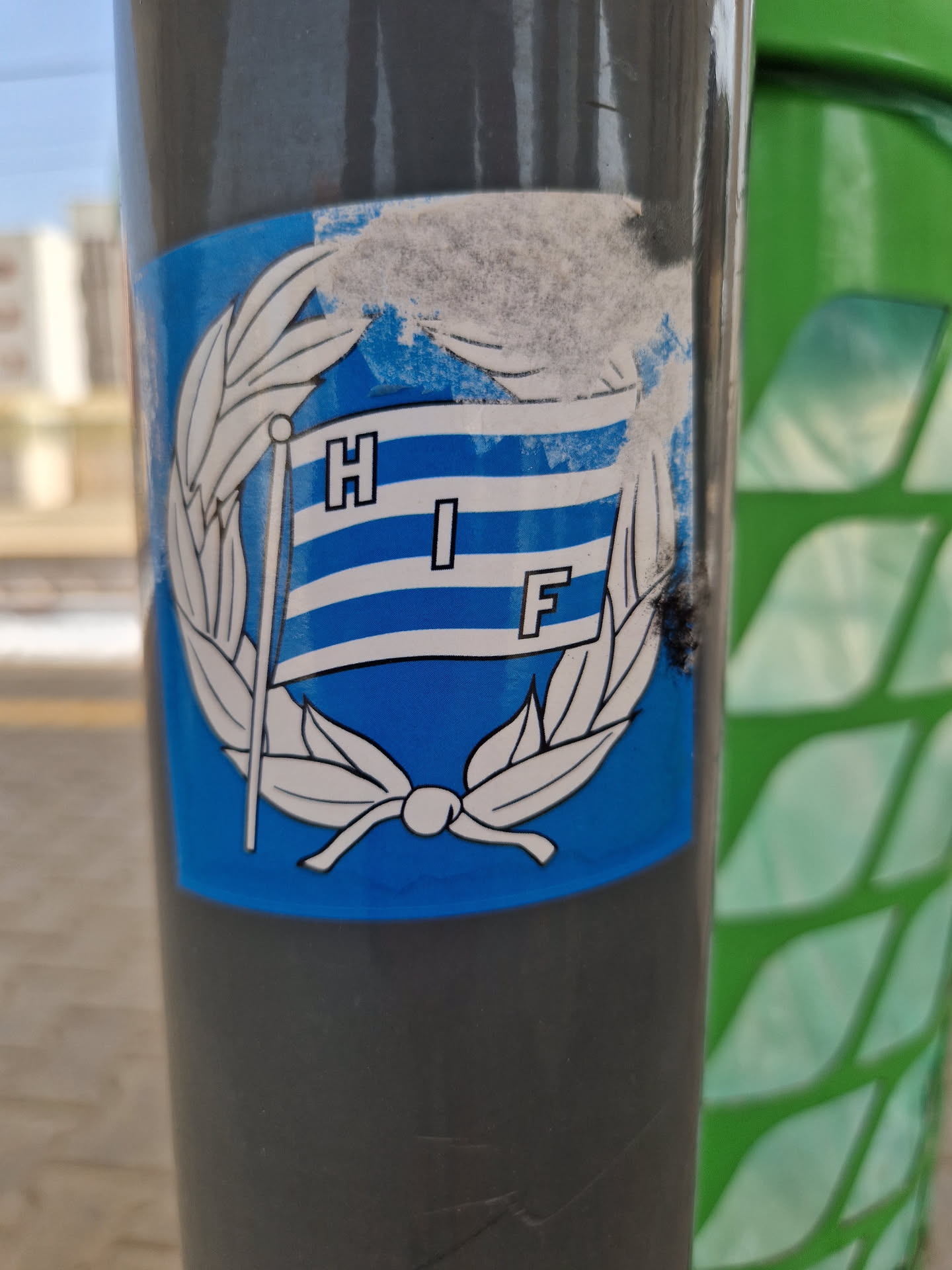 A blue sticker featuring a stylized flag with the letters HIF on it, surrounded by a laurel wreath. The sticker is affixed to a grey pole. The flag is blue and white striped, with the letters in a sans-serif font. The sticker shows some signs of wear and tear.