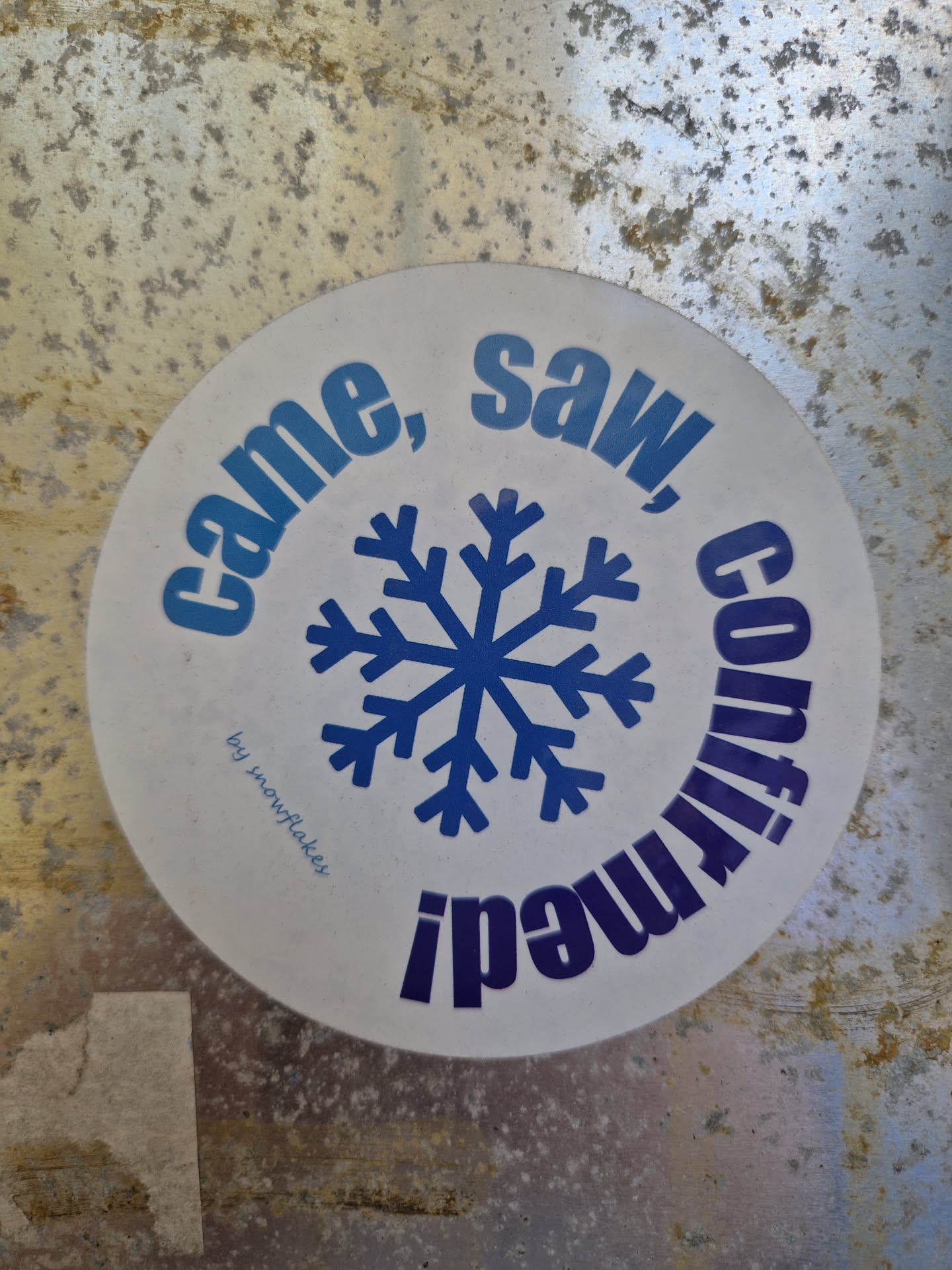 Round white sticker with a blue snowflake in the center and the text "Came, saw, conquered" written around it. The text is in a circular design and the words are in blue and purple.  The phrase "by snowflakes" is written in small text below the snowflake.