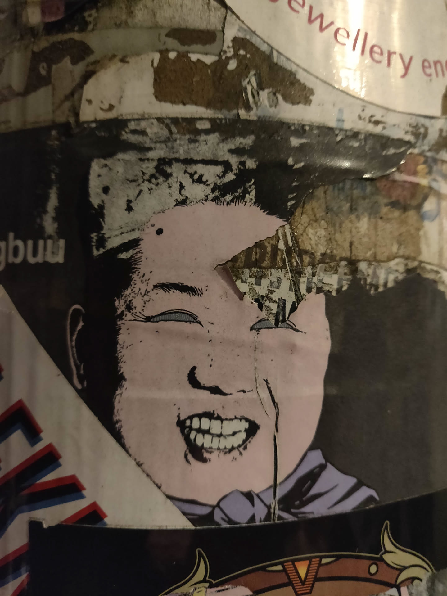 A weathered sticker depicting a caricature of Kim Jong Un with a wide, toothy grin.  The sticker shows some signs of wear and tear, suggesting it's been exposed to the elements for a while.