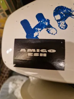 Black rectangular sticker with white text that says AMIGOESH. The sticker is placed on a white surface with blue graffiti in the background.