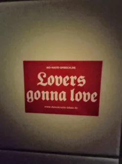 A red square sticker with white text.  The text says "Lovers gonna love" in a stylized font.  There are also smaller text at the top and bottom. The top text says "no-hate-speech.de" and the bottom says "www.demokratie-leben.de"