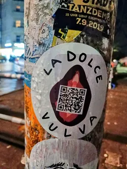 A round sticker with the phrase "La Dolce Vulva" written around a central image of a vulva with a QR code. The sticker is affixed to a pole covered in other stickers and graffiti. The QR code may link to a website or social media.