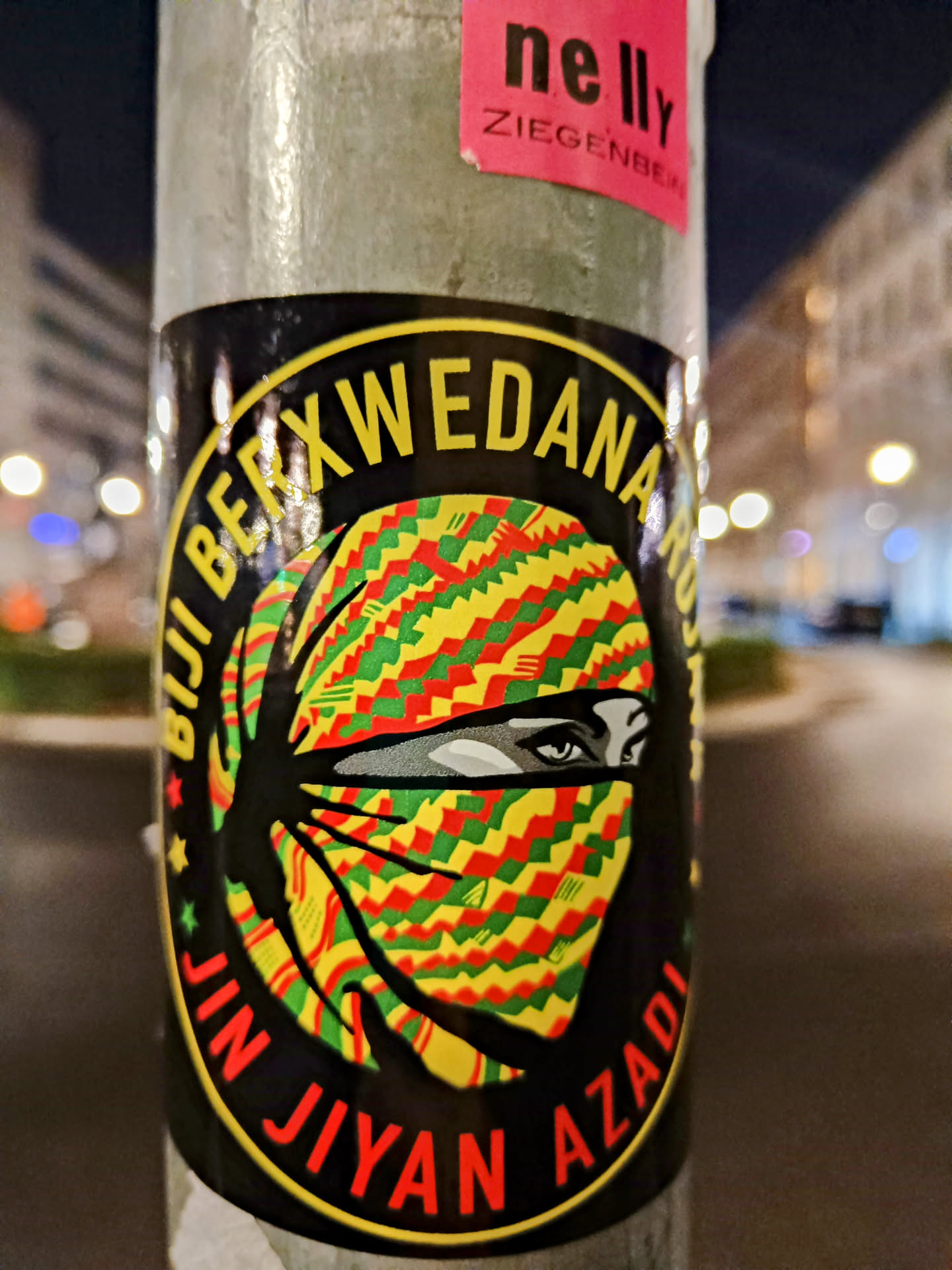 A round sticker featuring a woman's face partially covered by a patterned headscarf in yellow, green, and red. The text "Jin Jiyan Azadi" is written around the central image. The sticker is adhered to a pole.