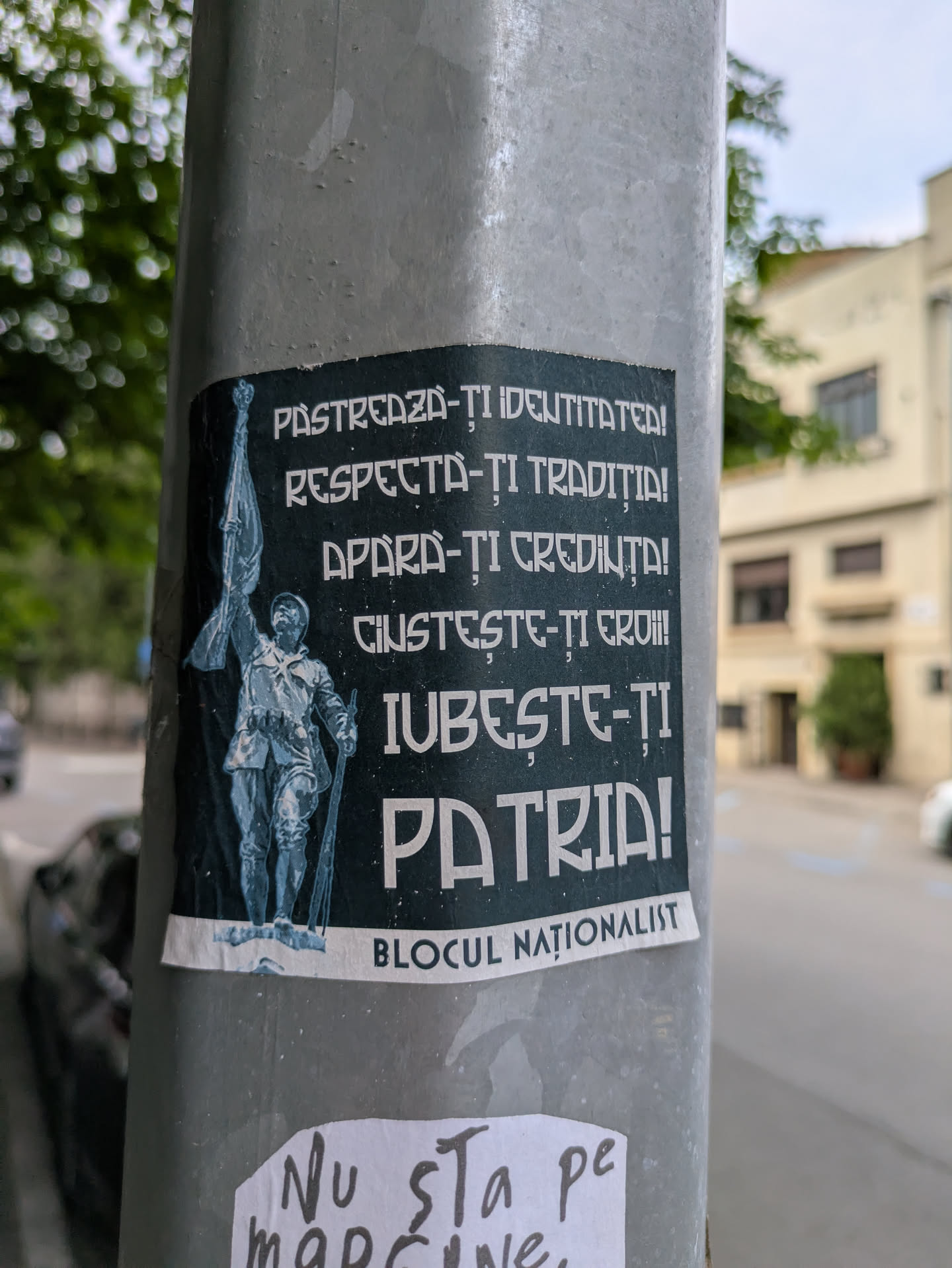 A sticker on a light pole features a nationalist message in Romanian. The text encourages people to preserve their identity, respect tradition, defend their faith, honor their heroes, and love their country. The sticker includes an illustration of a soldier with a flag.