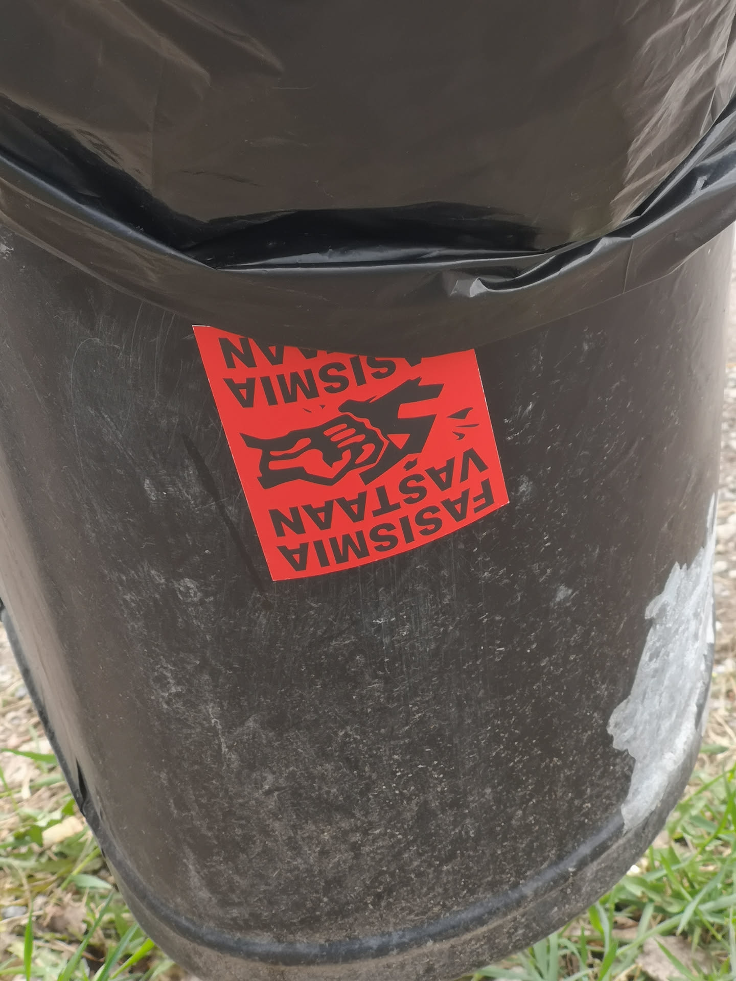 The sticker is rectangular with a vibrant red background. In the center, a stylized black graphic depicts a clenched fist or hand crushing a symbol, which is not fully discernible but implies a fascist emblem. The text 'FASISMIA VASTAAN' (Finnish for 'AGAINST FASCISM') is printed in black capital letters, appearing at both the top and bottom of the sticker, with the bottom text rotated 180 degrees relative to the top text. The sticker is adhered to a dark, textured surface, likely a trash bin, and shows some signs of wear and slight peeling at the edges.