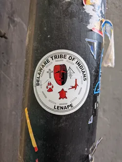 A circular sticker displaying the official seal of the Delaware Tribe of Indians (Lenape) adhered to a dark metal pole. The central motif is a stylized Mesingw face mask split vertically in red and black, surrounded by several cultural symbols: a cross, a traditional pump drill, a red turkey track, a red turtle, and a red wolf paw print representing clan animals. The outer rim contains bold black text reading 'DELAWARE TRIBE OF INDIANS' arched across the top and 'LENAPE' centered at the bottom, set against a white background.