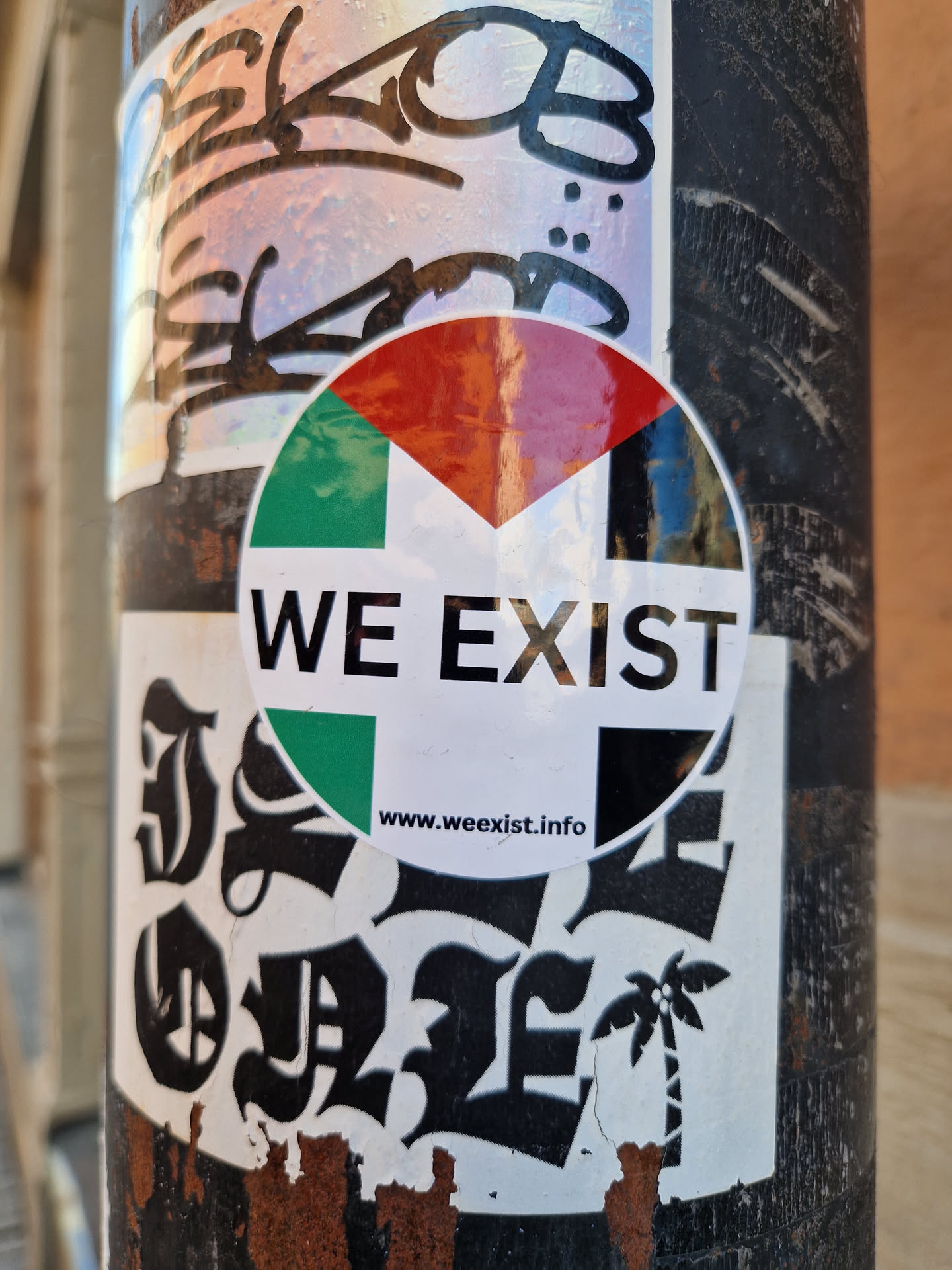 A round sticker with the text "WE EXIST" and a flag-like design in the background.  The bottom of the sticker has a URL and a small palm tree graphic. It's affixed to a metal pole with other stickers.