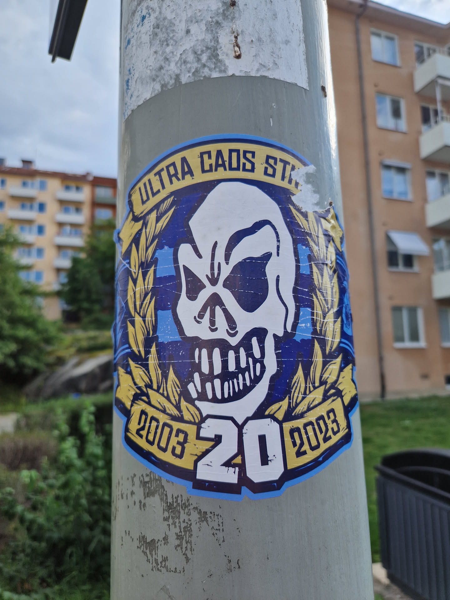 A sticker featuring a skull graphic with text that reads "ULTRA CAOS STAND 2003 20 2023". The skull is encircled by a laurel wreath-like design. The color scheme is primarily blue, yellow, and white.