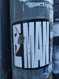 Black and white sticker with the word "LIMAN" stylized with bold lettering and a small angel with 666 written above it. The sticker is applied to a metallic pole. There are some small black spots on the sticker.