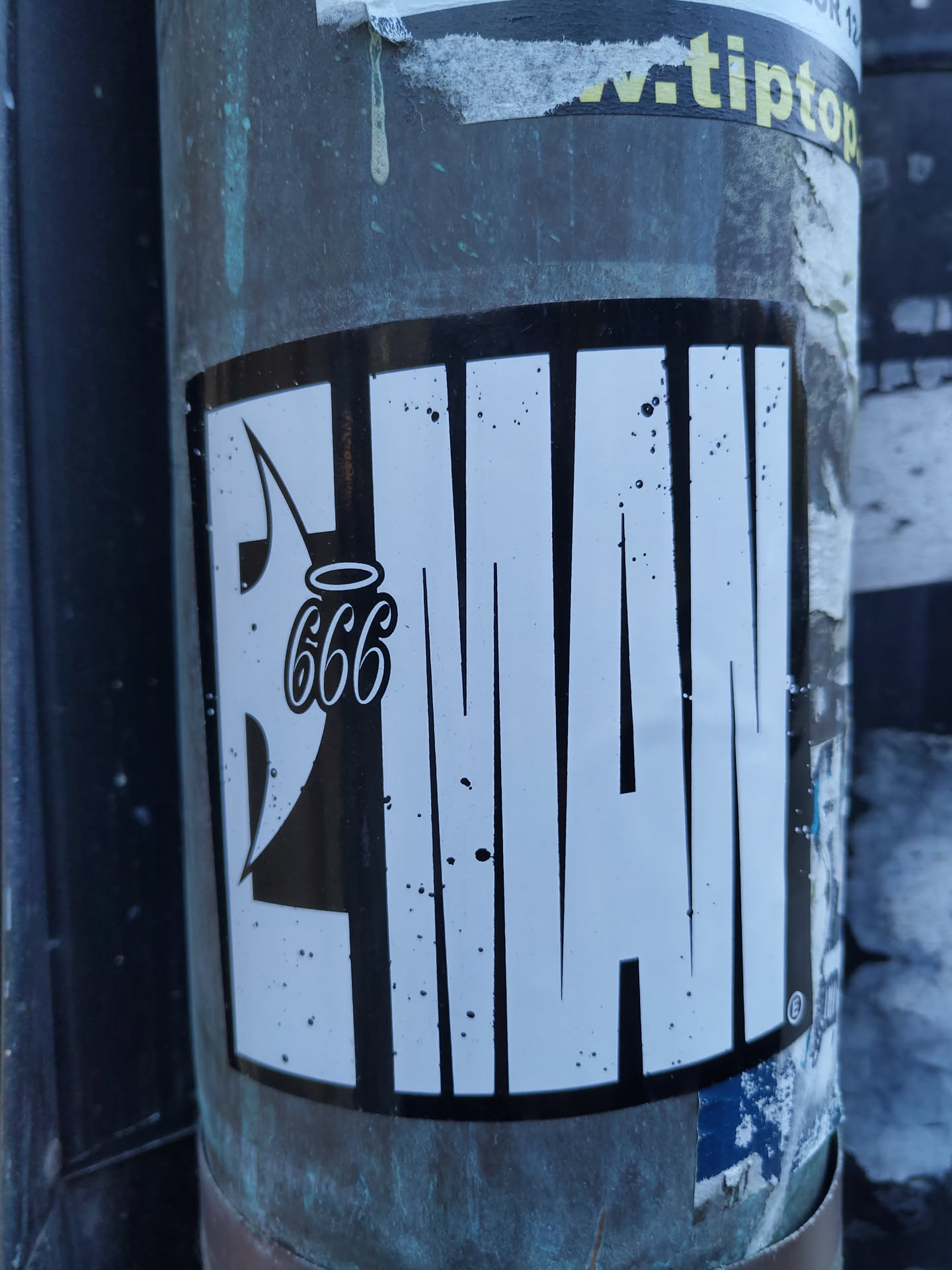 Black and white sticker with the word "LIMAN" stylized with bold lettering and a small angel with 666 written above it. The sticker is applied to a metallic pole. There are some small black spots on the sticker.
