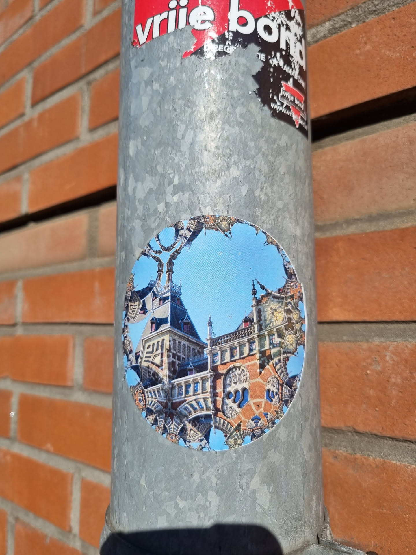 This round sticker is affixed to a metal pole. The main image appears to be a photograph of a building, possibly in Amsterdam, with an artistic distortion or kaleidoscopic effect. The colors are vibrant, and the building's details are visible despite the altered perspective. The sticker itself has a slightly worn appearance, suggesting some time spent outdoors.