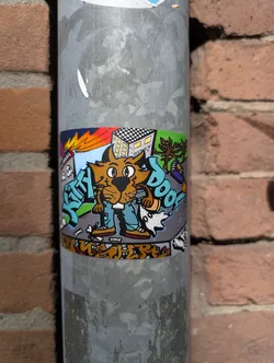 This sticker features a cartoon cat character in jeans and shoes, surrounded by graffiti-style elements, including buildings, a tree, and dog bones. The background includes a building with a checkered roof and some graffiti, adding to its urban art style. The sticker is attached to a metal pole.