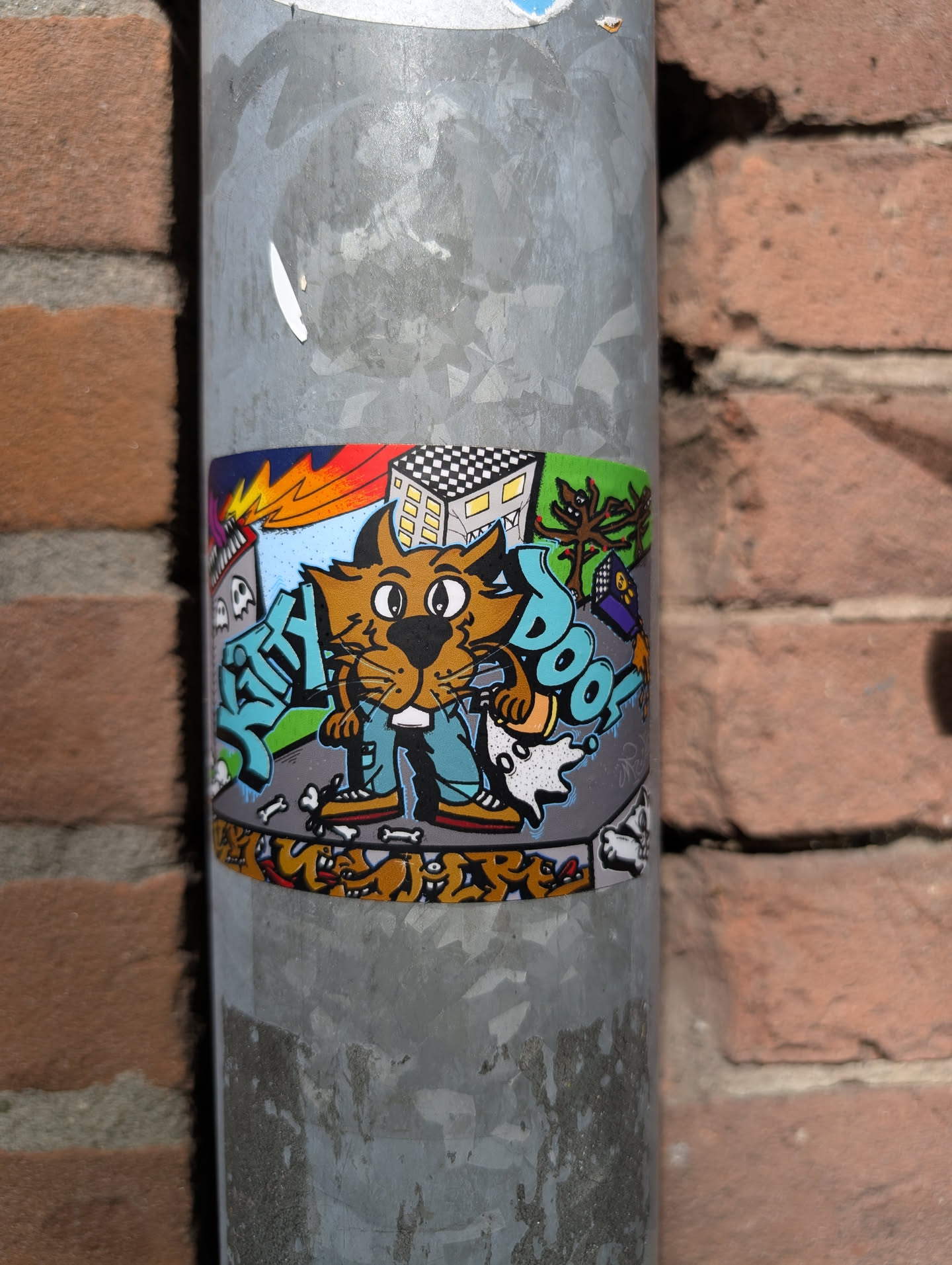 This sticker features a cartoon cat character in jeans and shoes, surrounded by graffiti-style elements, including buildings, a tree, and dog bones. The background includes a building with a checkered roof and some graffiti, adding to its urban art style. The sticker is attached to a metal pole.