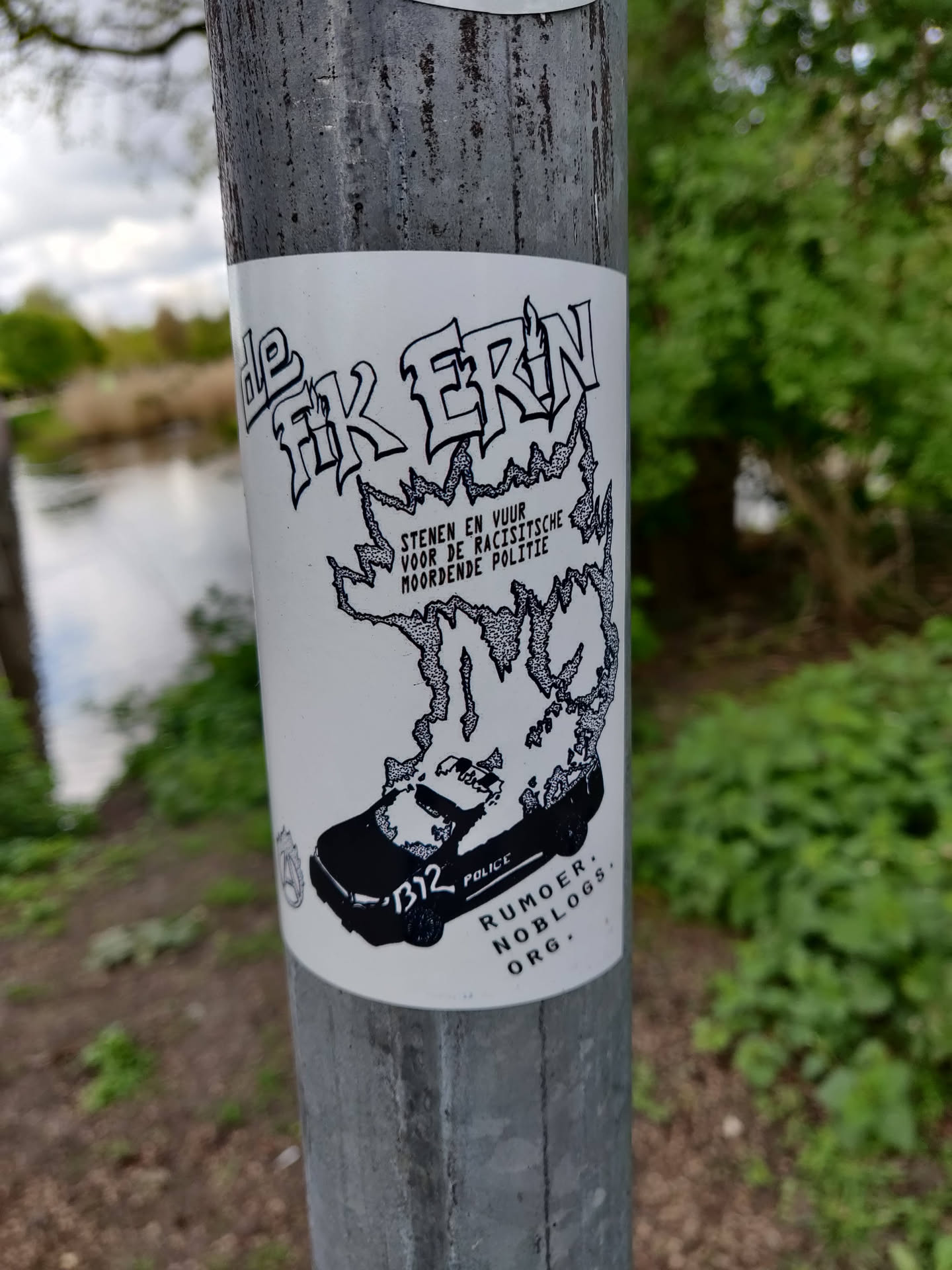 A sticker featuring a black and white illustration of a police car engulfed in flames.  The Dutch text translates roughly to "set it on fire" in relation to racist and murderous police. The bottom features a website address: rumboer.noblogs.org. The style is reminiscent of protest art.