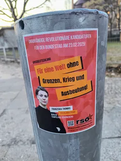A red political sticker affixed to a metal pole, advertising "Unabhängige Revolutionäre Kandidaturen" for the Bundestag election on February 23, 2025. The sticker features a photo of Franziska Thomas, identified as a social worker from Berlin-Tempelhof-Schöneberg. It includes the slogan "Für eine Welt ohne Grenzen, Krieg und Ausbeutung!" (For a world without borders, war and exploitation!).