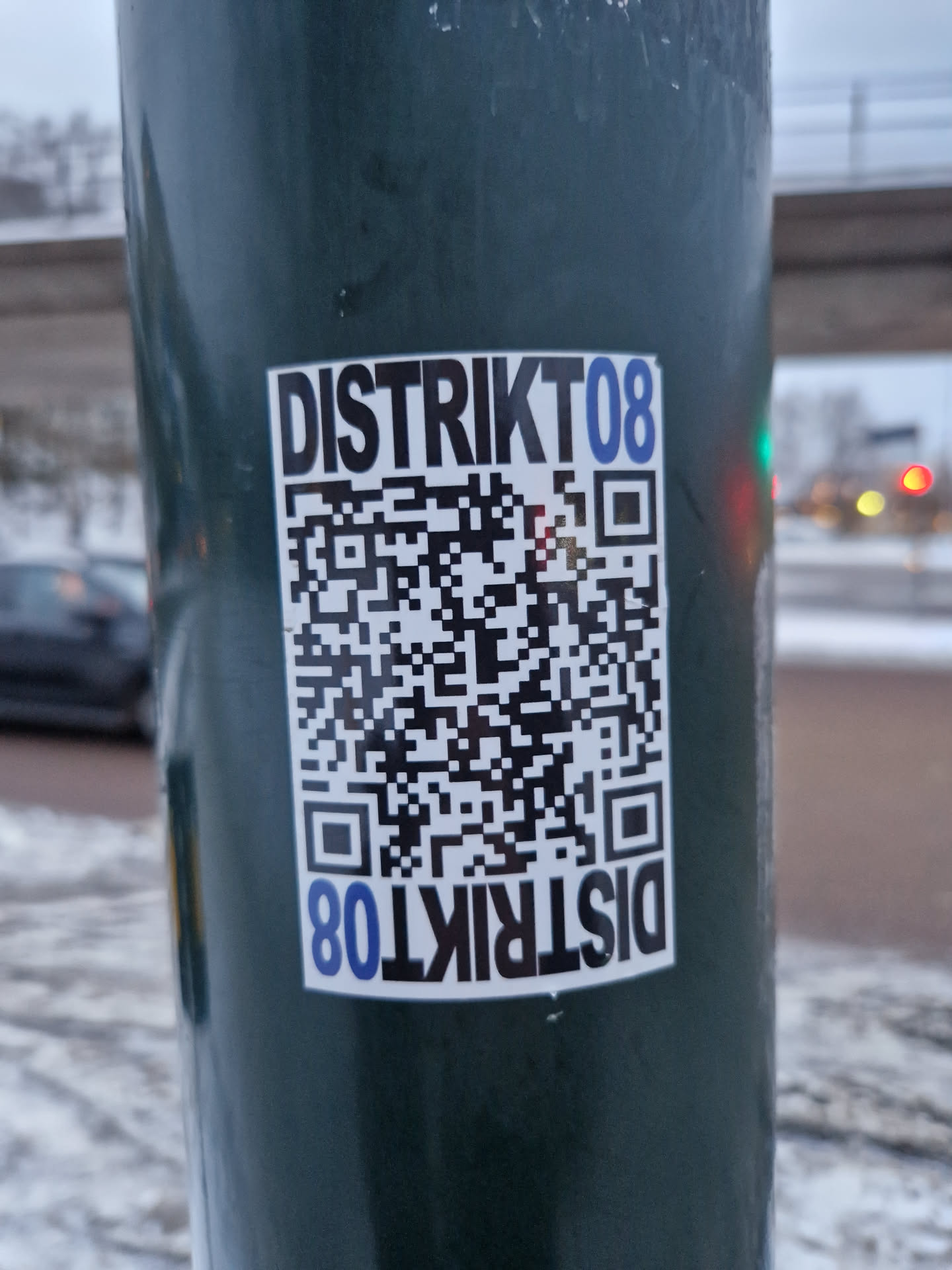 A sticker featuring the text 'Distrikt08' and a QR code is affixed to a green pole.  The QR code appears to link to a website or social media page. The sticker is rectangular and printed on white paper.
