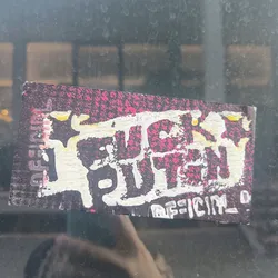 A sticker with the phrase "FUCK PUTIN" in stylized graffiti lettering. The background is dark with pink splatter. The sticker seems to be handmade or DIY style.
