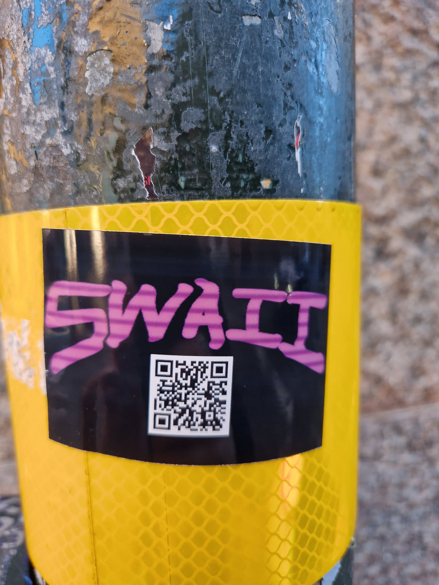 A black sticker with the word "SWaii" written in purple and a QR code underneath. The sticker is placed on a yellow reflective surface.